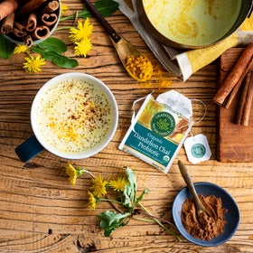 A cup of Dandelion Chai Probiotic Tea on a wooden table, accompanied by spices, turmeric, and a teabag, supports healthy digestion.