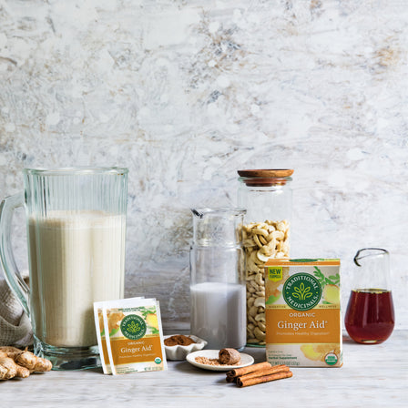 Ginger Aid® Tea, renowned for supporting healthy digestion, is placed next to a pitcher of smoothie, nuts, and spices arranged on a marble surface.