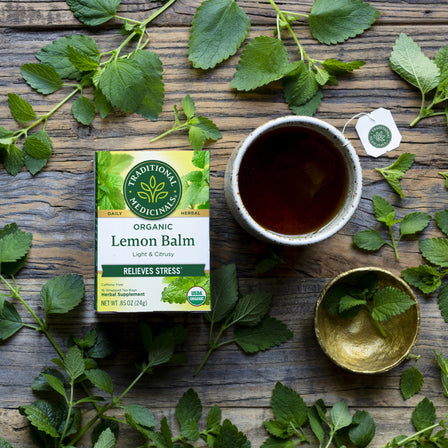 On a wooden table sits a box of Lemon Balm Tea, next to a steaming cup and surrounded by fresh lemon balm leaves. This calming tea soothes the nervous system and aids digestion, providing tranquility with every sip.