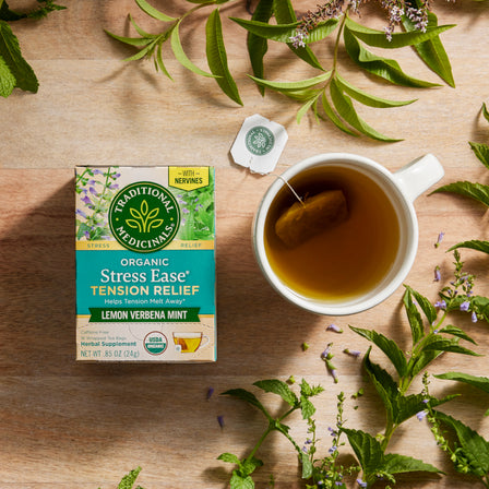 A box of Stress Ease® Tension Relief Tea is placed next to a steaming cup, encircled by fresh green leaves and a touch of skullcap on a rustic wooden surface, exuding relaxation.